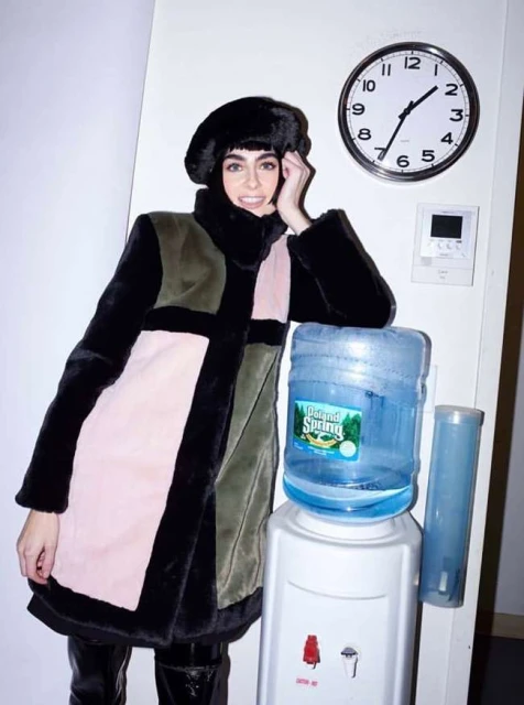 Woman in fur coat and hat stands next to a water cooler with a clock and thermostat visible on the wall in an indoor setting.
