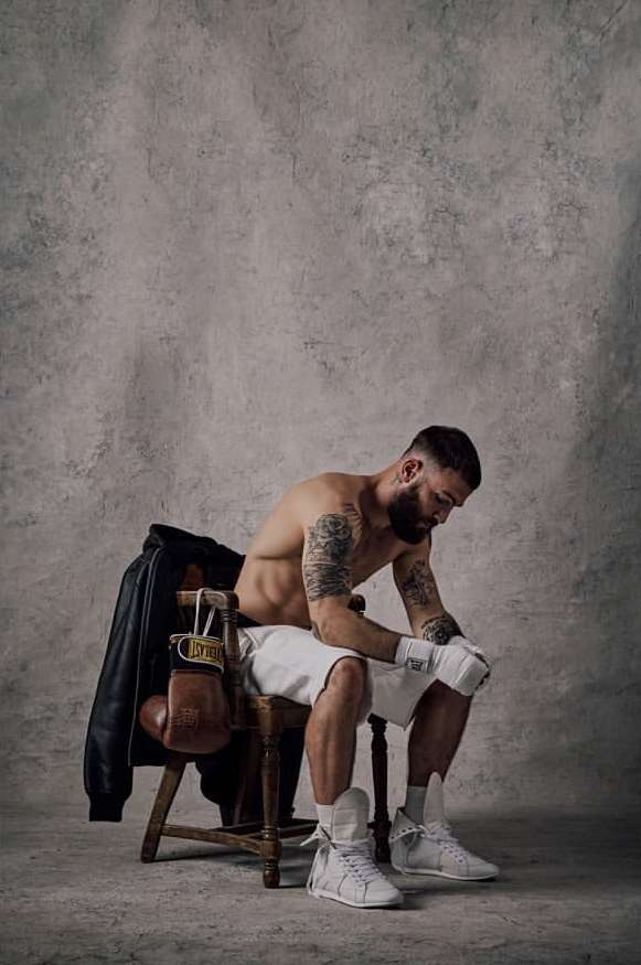 Tattooed male boxer with wraps sitting on a chair with boxing gloves and a black jacket in an indoor studio setting.