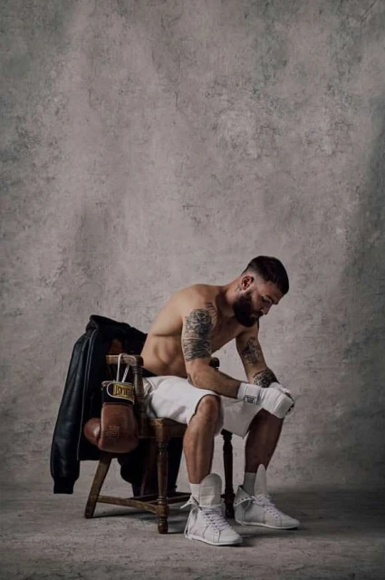 Tattooed male boxer with wraps sitting on a chair with boxing gloves and a black jacket in an indoor studio setting.