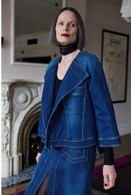 Elegant woman in denim jacket and skirt, posing indoors. Features include short gray hair, black top and scarf, fireplace, and artwork.
