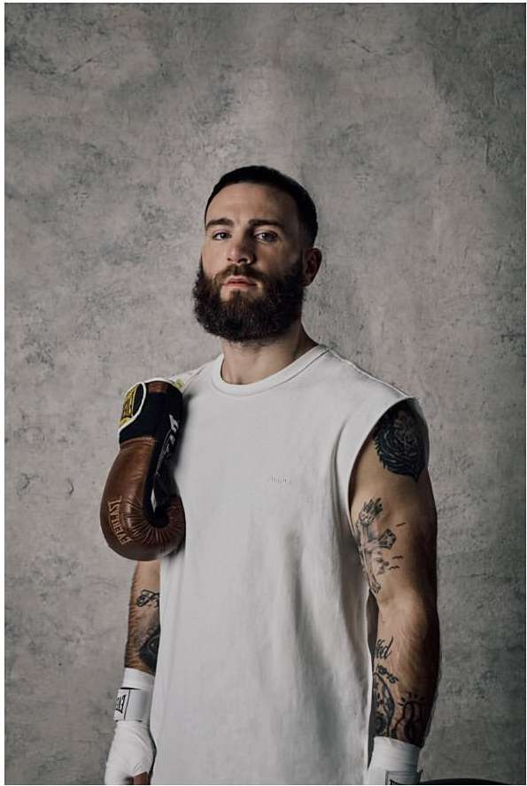 Portrait of a bearded man with tattoos and boxing gloves, wearing a white t-shirt, posed in front of a textured gray background.