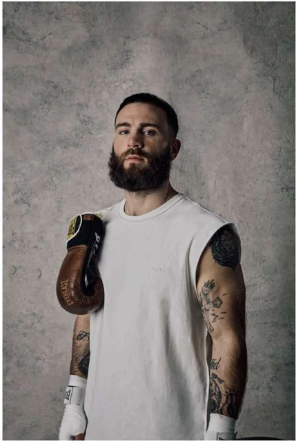 Portrait of a bearded man with tattoos and boxing gloves, wearing a white t-shirt, posed in front of a textured gray background.