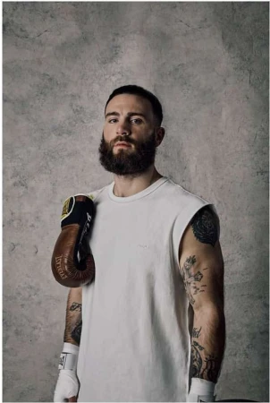 Portrait of a bearded man with tattoos and boxing gloves, wearing a white t-shirt, posed in front of a textured gray background.