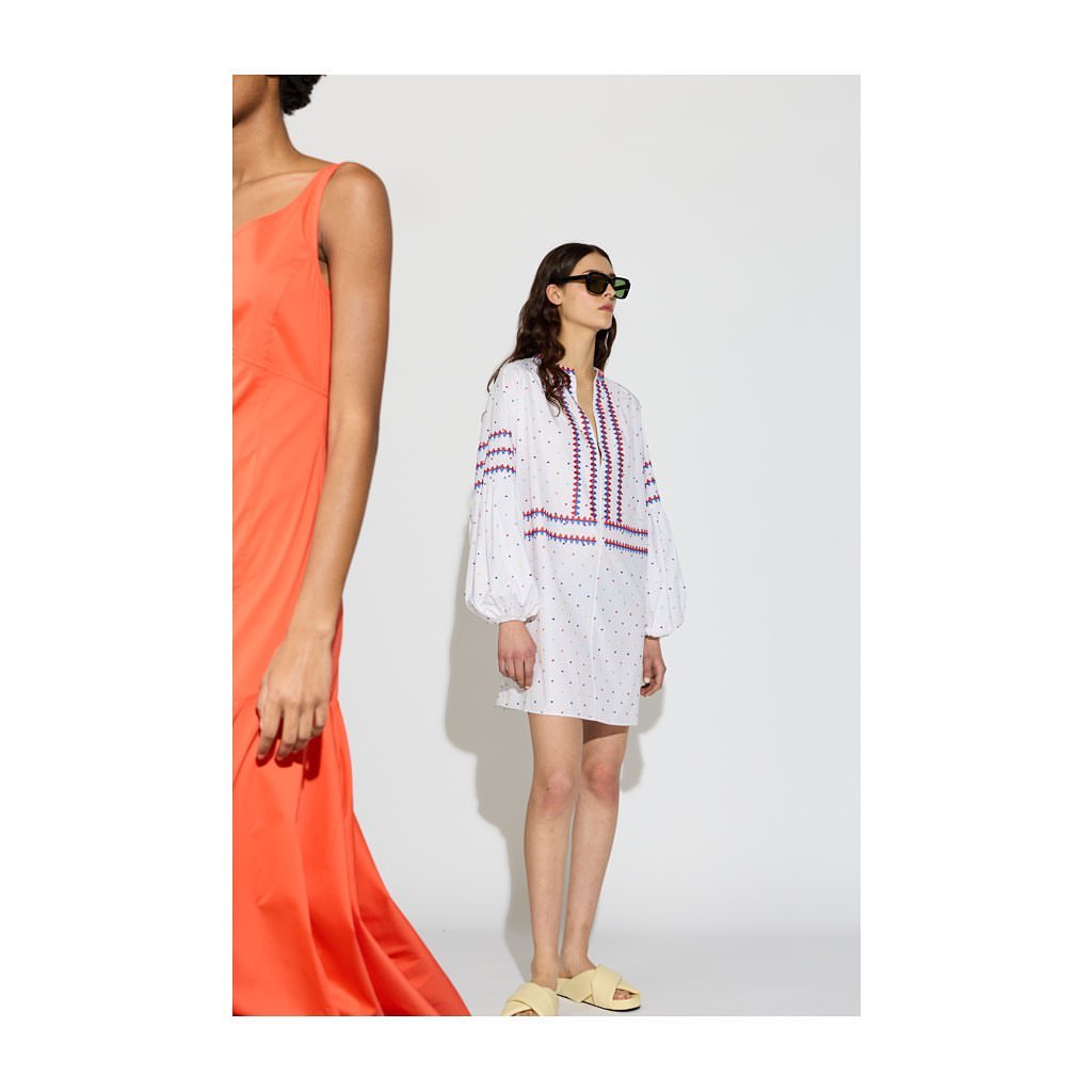 Two women stand in a brightly lit studio, one in a coral dress, the other in a white dress with colorful embroidery and wearing sunglasses.