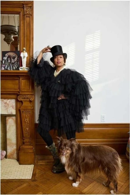 Adult female in a black ruffled coat and hat poses indoors with a brown dog near a fireplace; light wall and wood trim.