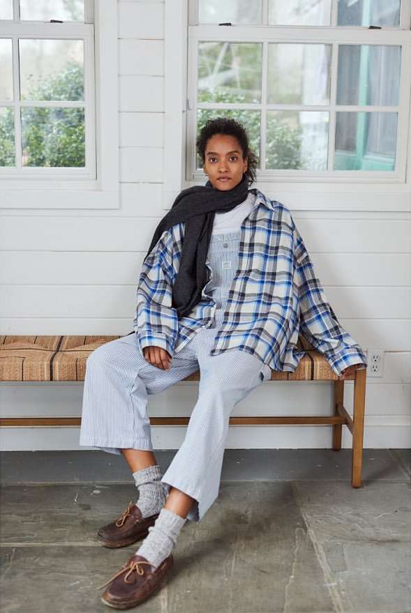 Adult female sitting on a bench in a flannel shirt and scarf indoors near a window, wearing striped overalls and leather loafers.