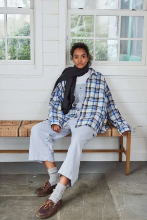 Adult female sitting on a bench in a flannel shirt and scarf indoors near a window, wearing striped overalls and leather loafers.