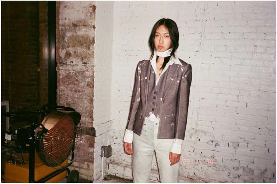 Fashion portrait of a young adult female in a grey blazer and white pants, posing in front of a distressed brick wall in a studio.
