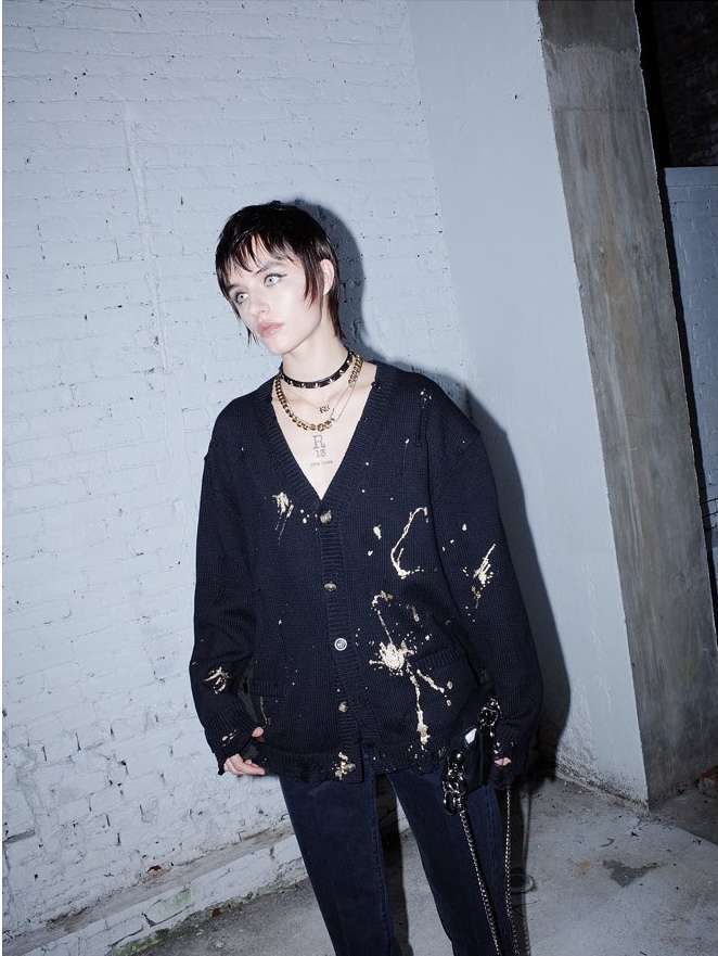 Woman with short dark hair posing in a distressed black cardigan and dark jeans against a neutral brick wall with natural lighting.