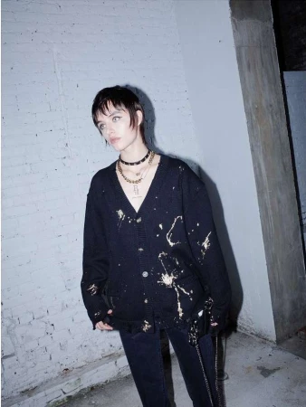 Woman with short dark hair posing in a distressed black cardigan and dark jeans against a neutral brick wall with natural lighting.