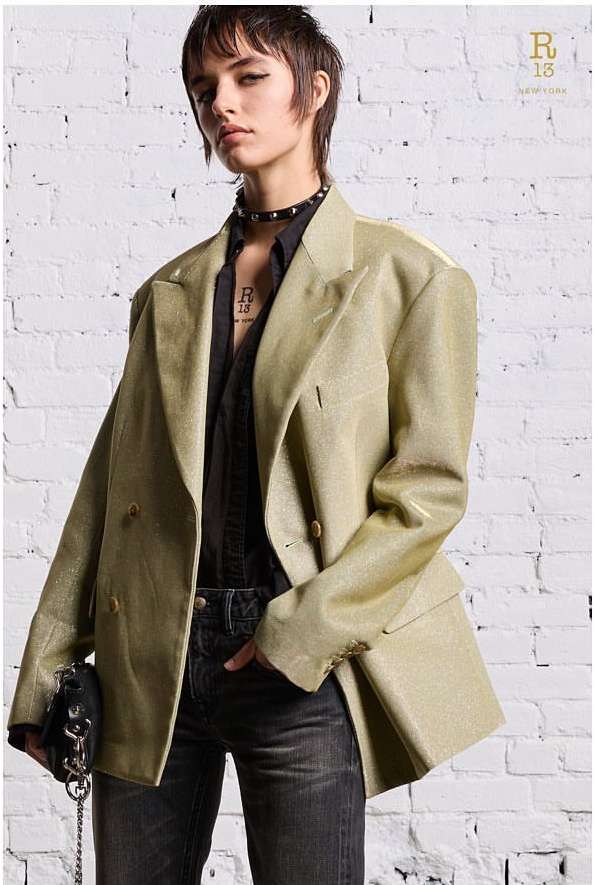 Fashion shot of a woman wearing a light-gold, oversized blazer with black top, jeans and studded choker against a white brick wall.