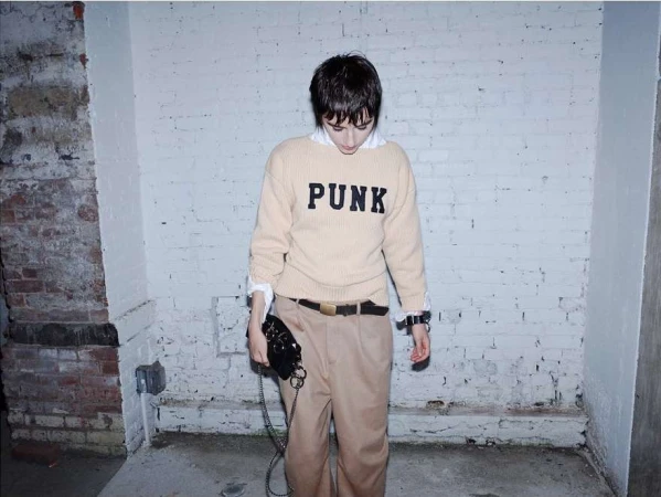 Model wearing a beige sweater with the word PUNK printed across the chest against a white brick wall.