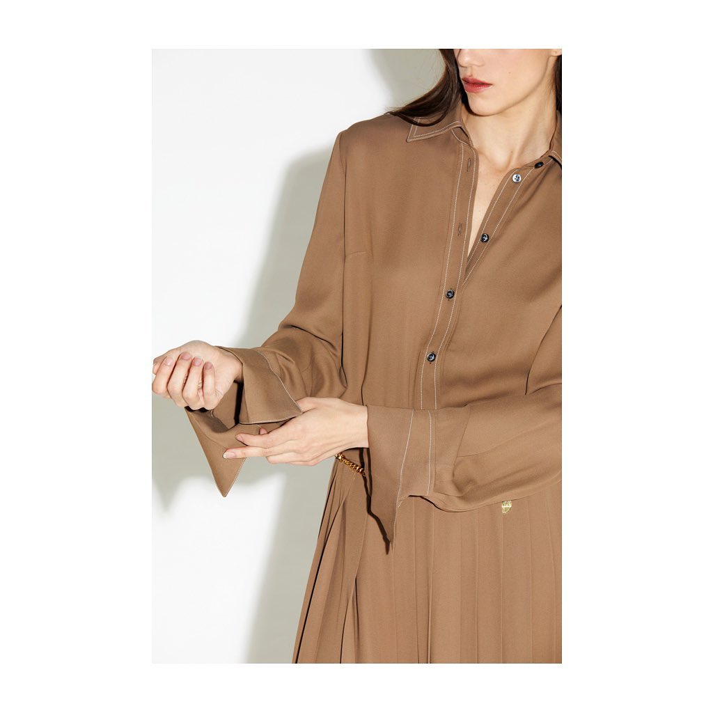 Woman in a light brown shirt dress and gold chain belt poses in a studio with a white backdrop. Fashion shoot.