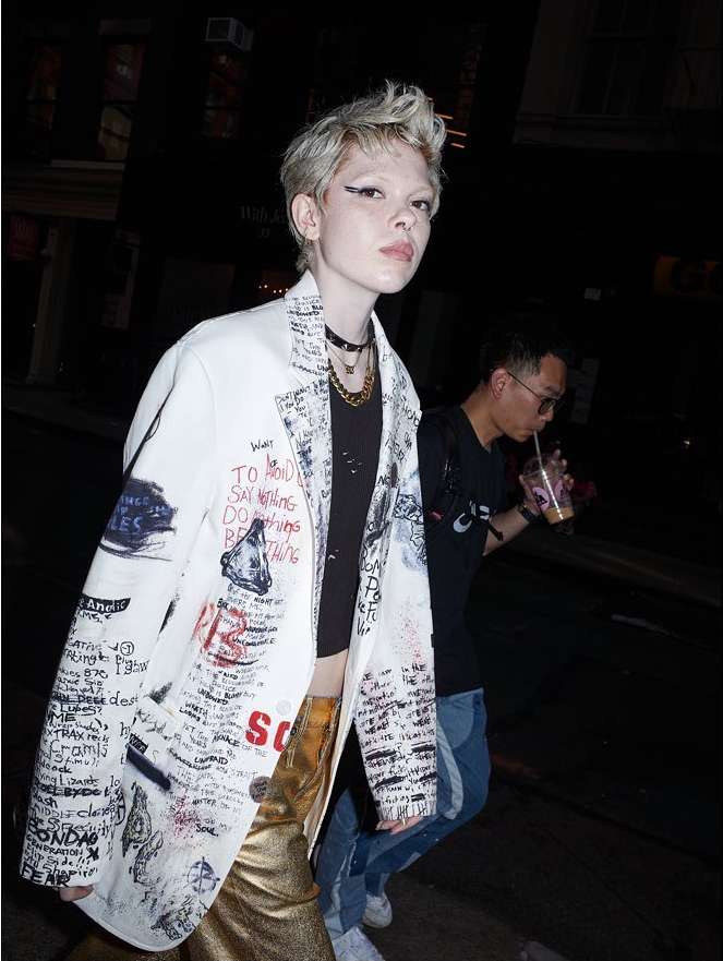 A person wearing a blazer with handwritten text, gold pants, and multiple necklaces stands in a dimly lit urban street at night.