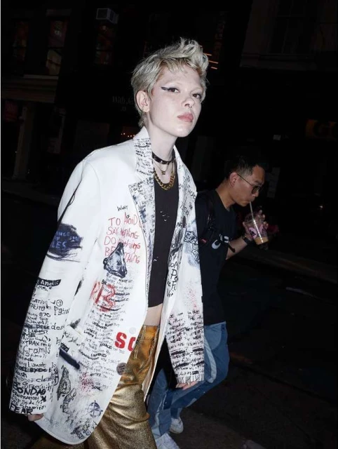 A person wearing a blazer with handwritten text, gold pants, and multiple necklaces stands in a dimly lit urban street at night.