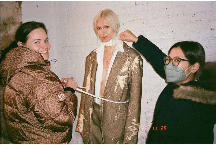 Three people indoors, one wearing a mask, one wearing a leopard print jacket, and one getting measured with brown, painted-style jacket.