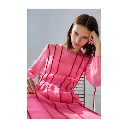 Woman in a pink dress with red vertical stripes seated on a metal chair against a plain white wall