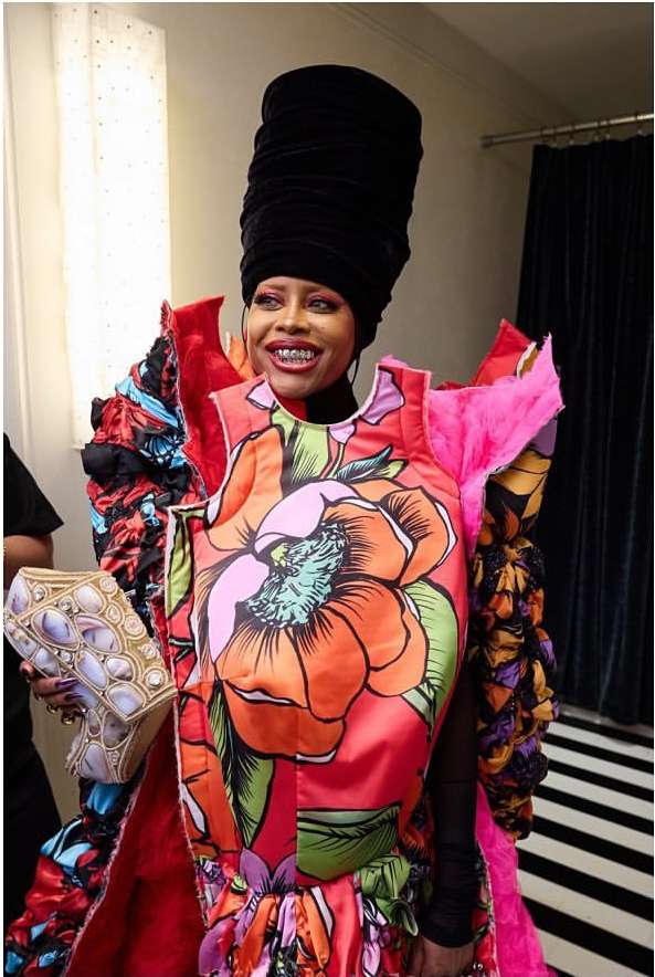 An adult female smiles while wearing a high-fashion floral dress and a tall black head wrap in an indoor environment. She is also carrying a jeweled purse.