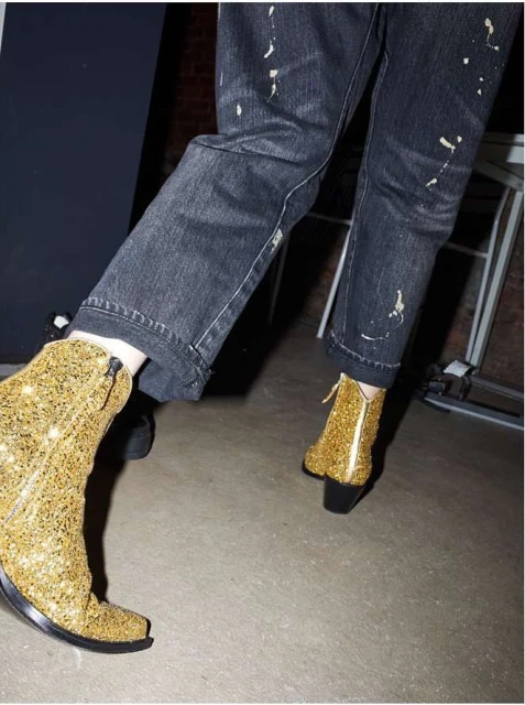 Close-up of a person wearing sparkly, gold, ankle boots and paint-splattered, black jeans in an indoor setting with neutral background.