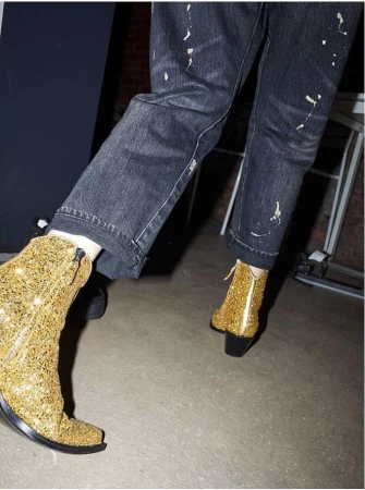 Close-up of a person wearing sparkly, gold, ankle boots and paint-splattered, black jeans in an indoor setting with neutral background.
