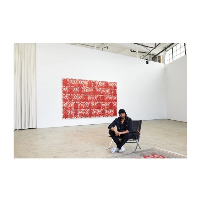 Man sits in a modern chair in a large, light-filled white-walled gallery space, with red patterned artwork behind him, and carpet nearby.