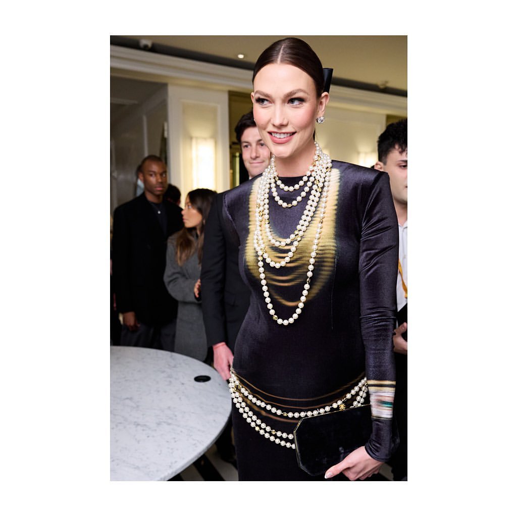Woman in black dress adorned with multiple strands of pearls, posing indoors with other people in the background.