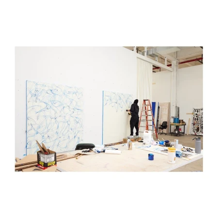 Artist in black hooded jumpsuit stands on ladder in studio, painting blue lines on white canvas, art supplies and materials visible.