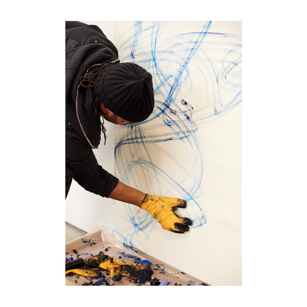 Artist creating abstract drawing on white wall using blue pastel; yellow work gloves and tray with pastel debris visible.