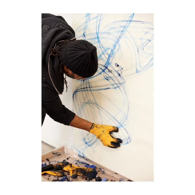 Artist creating abstract drawing on white wall using blue pastel; yellow work gloves and tray with pastel debris visible.