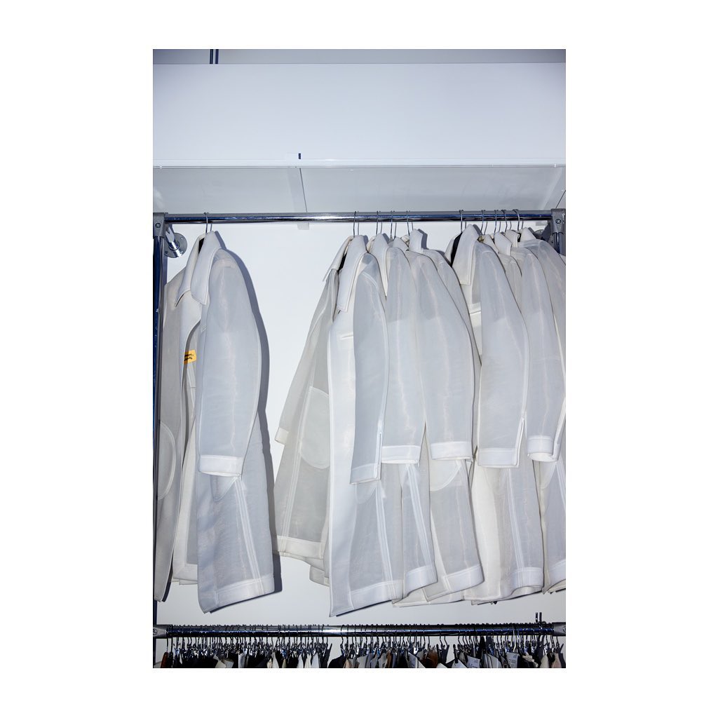 Rack of white lab coats or dust coats on hangers under fluorescent lighting, ready for use in a medical or cleanroom setting.