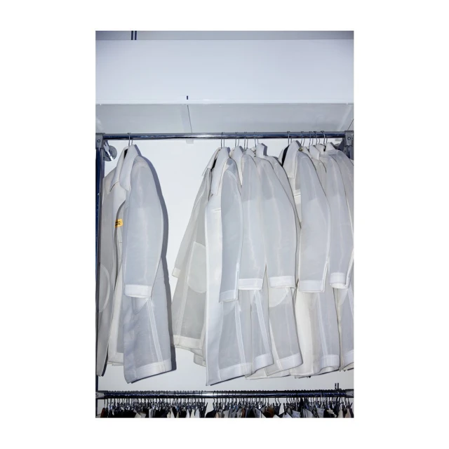Rack of white lab coats or dust coats on hangers under fluorescent lighting, ready for use in a medical or cleanroom setting.