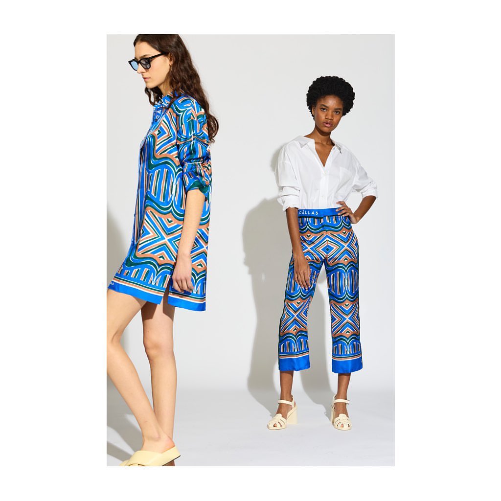 Two fashion models pose in a bright studio wearing vibrant blue patterned outfits, one in a dress and the other in a white shirt and pants.