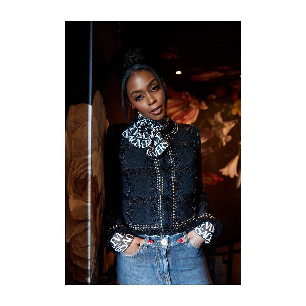 Portrait of an adult Black woman with her hair in a topknot, wearing a Versace-print scarf and jacket with denim pants indoors.
