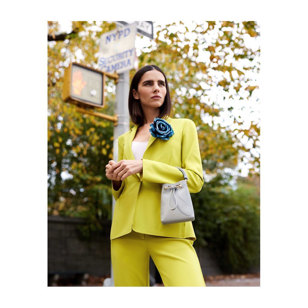 Woman in a chartreuse pantsuit stands in front of a street sign in an urban setting, holding a gray purse.