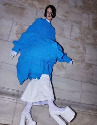 Model wearing an oversized bright blue sweater and white platform heels against a stone block background.