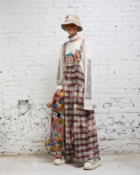 Model wearing a plaid overall and bucket hat holding a skateboard against a white painted brick wall