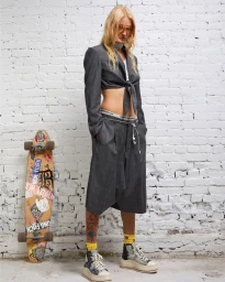 Model with blonde hair wearing grey cropped blazer and trousers standing next to a sticker-covered skateboard against a white brick wall