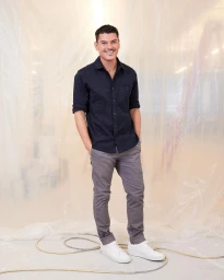 Man wearing a black buttoned shirt, grey trousers, and white sneakers posing in front of a wrinkled plastic backdrop