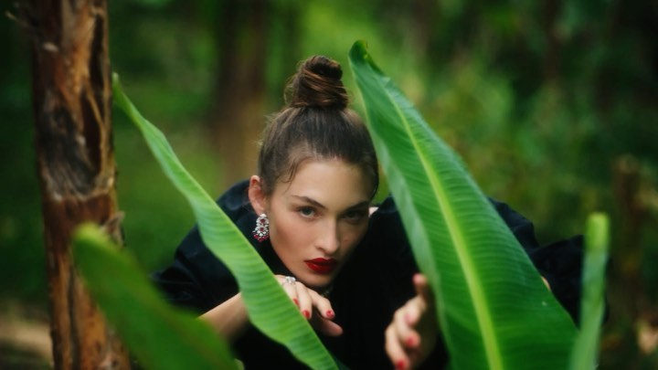 Portrait of a brunette woman with her hair in a bun, red lipstick, and earrings, framed by large tropical leaves in an outdoor setting.