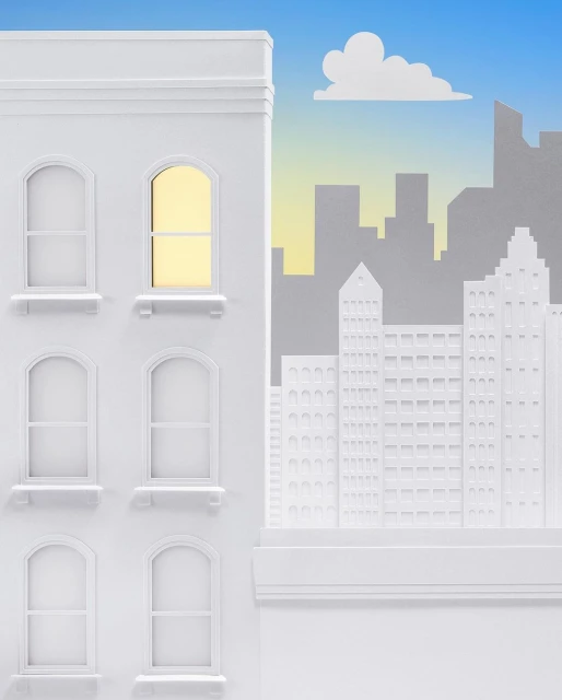 Paper art featuring a white building with lighted window against a cityscape silhouette and blue sky background, showcased in a studio setting.