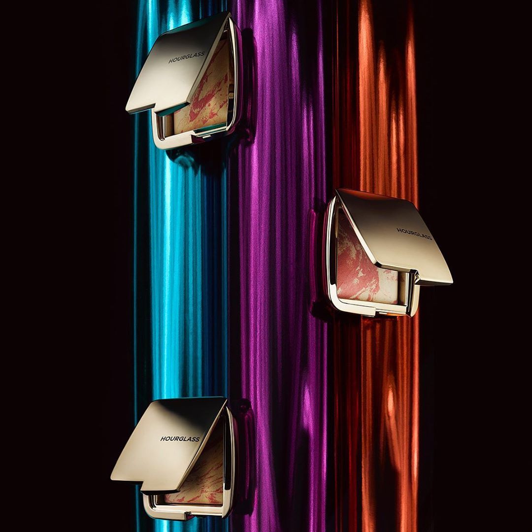 Three hourglass makeup compacts are displayed on a black surface with colorful vertical stripes in the background.