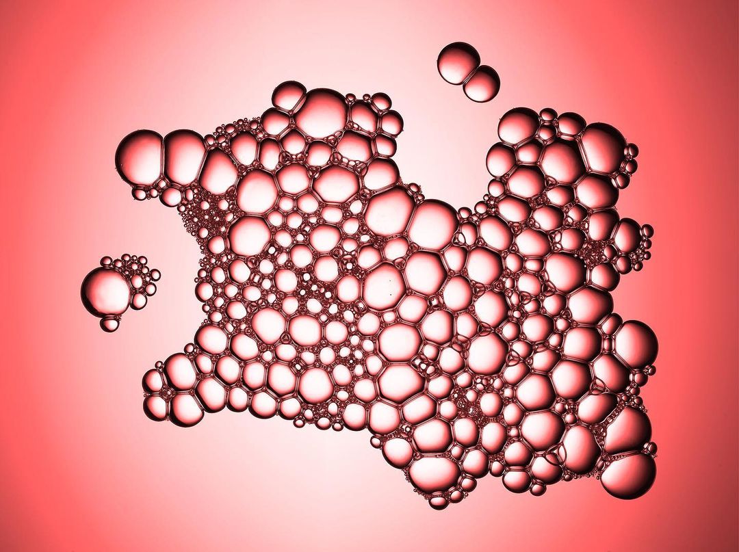 Macro photograph showing a collection of translucent red bubbles against a graduated red background with soft light, creating a cellular aesthetic.