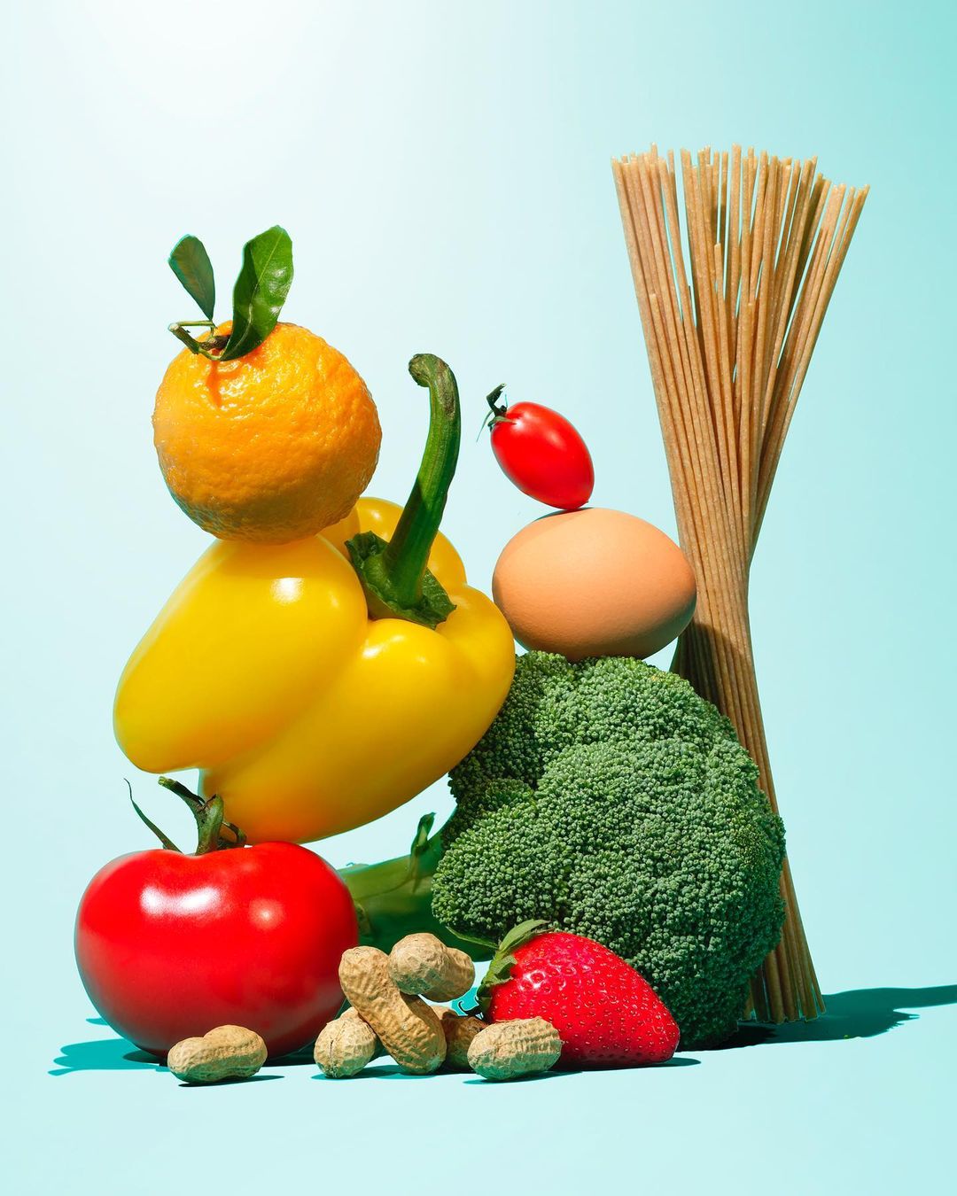 Still life composition featuring an arrangement of fresh produce including tomatoes, peppers, broccoli, pasta, peanuts, and an egg.
