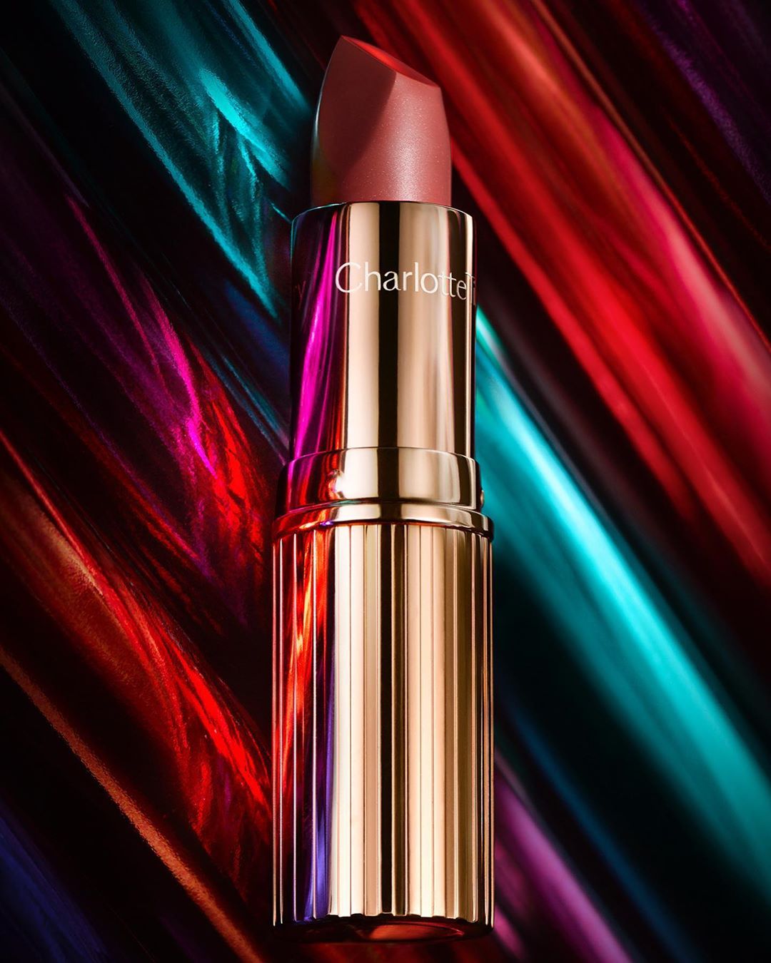 Close-up of a gold Charlotte Tilbury lipstick tube with a rose-colored lipstick bullet against a colorful light painted background.