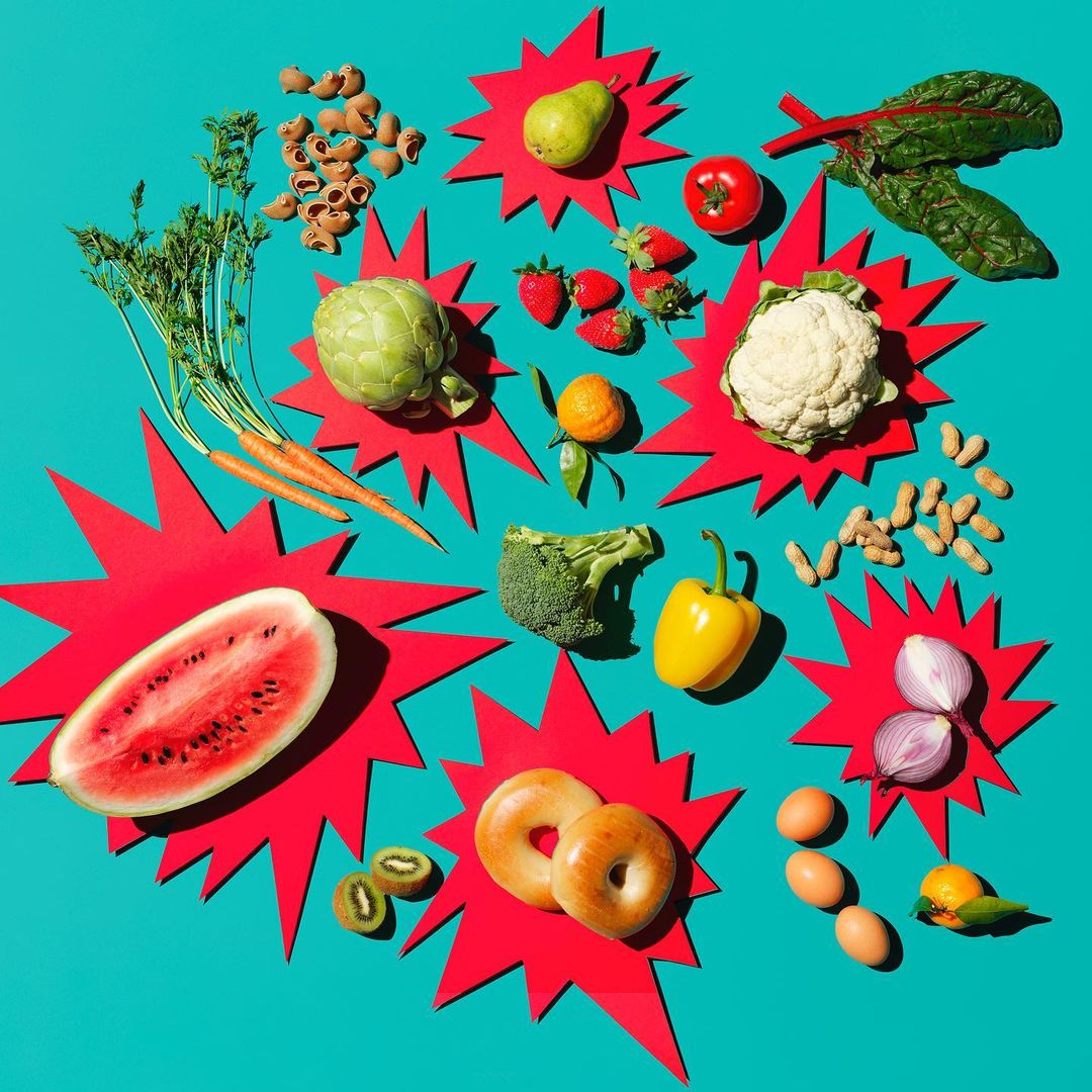Overhead shot of colorful fresh produce on a vibrant teal background, including watermelon, artichoke, bagels, and varied fruits and vegetables.