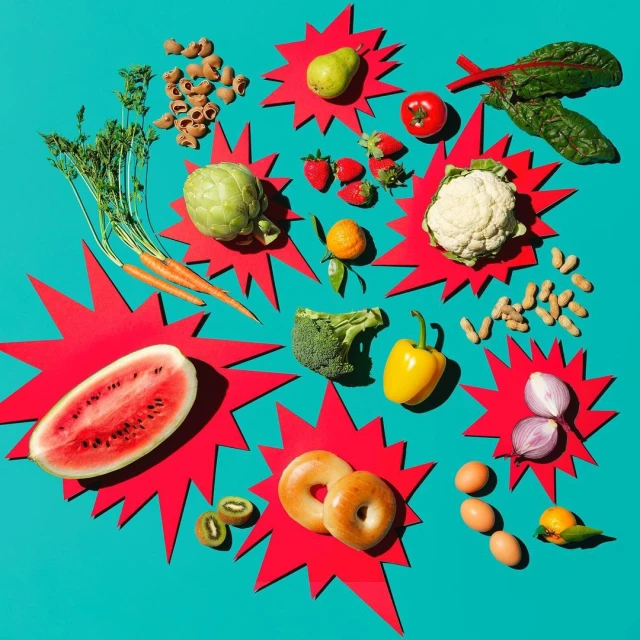 Overhead shot of colorful fresh produce on a vibrant teal background, including watermelon, artichoke, bagels, and varied fruits and vegetables.