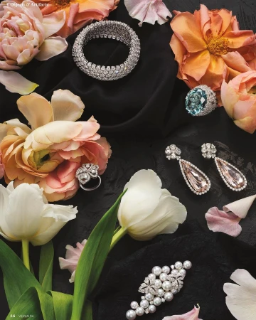 Still life of jewellery including a bracelet, earrings, and rings arranged with peach and white flowers on a black cloth background.