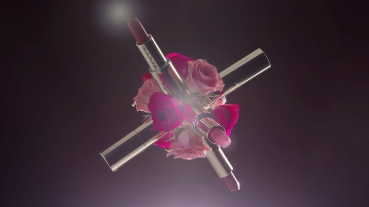 Cosmetic product: Three Becca brand lipsticks are crossed over pink roses against a soft dark background with light flares.