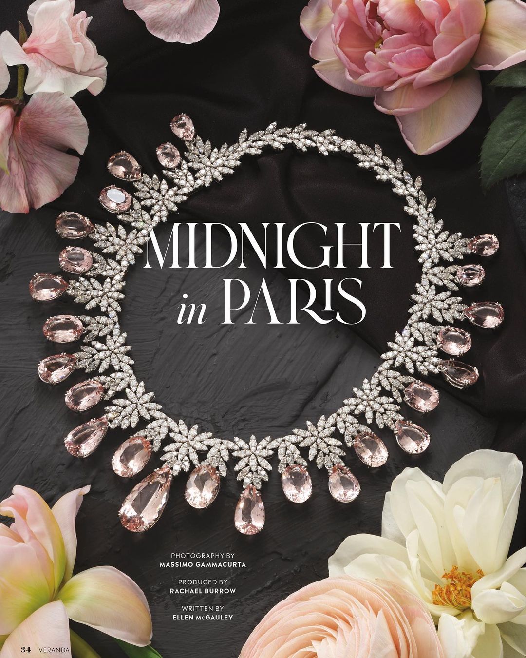 Close-up of a diamond and light pink gemstone necklace surrounded by pink and white flowers on a dark grey stone surface.