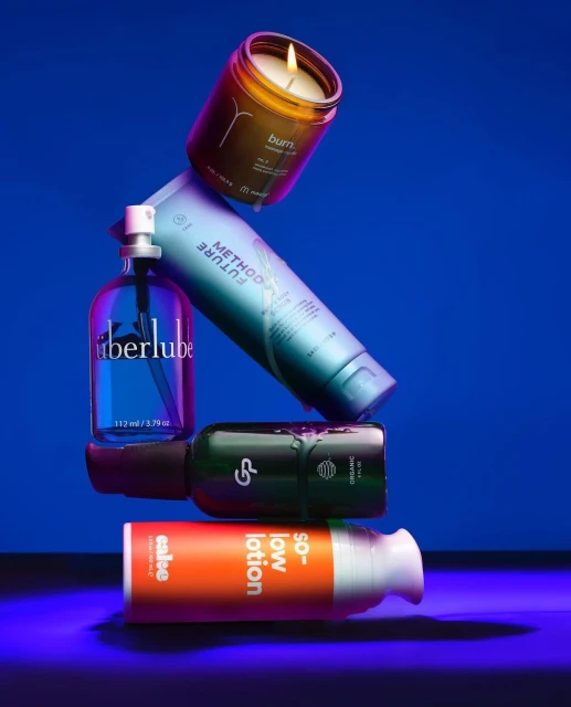 Stacked arrangement of beauty and personal care products including candle, lotion, and lubricant in a studio setting with blue background.
